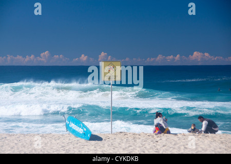 Dangerous Currents sign next to lifeguard surfboard with family playing on beach and stormy sea Bronte Sydney NSW Australia Stock Photo