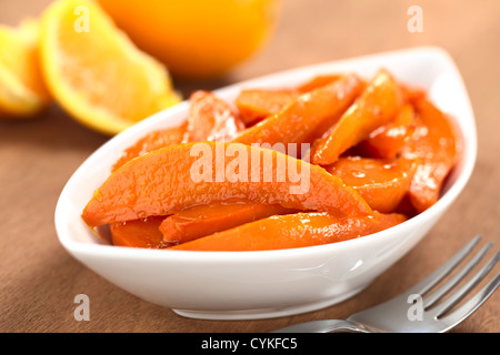 Bowl of sweet potato wedges caramelized with brown sugar and fresh orange juice with a fork on the side and oranges in the back Stock Photo