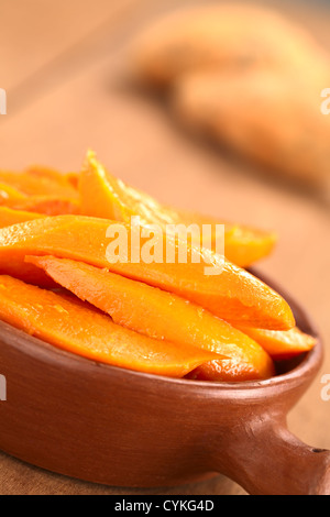 Sweet potato wedges caramelized with brown sugar and fresh orange juice in rustic bowl with sweet potatoes in the back Stock Photo