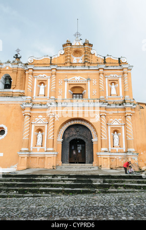 Iglesia San Pedro Las Huertas Colonial Architecture Guatemala | SAN PEDRO LAS HUERTAS, Guatemala — Viewed head-on from the cobblestone plaza, the Iglesia de San Pedro las Huertas shows its full ornamented facade, dated 1672, with white stucco statuary set against burnt-sienna walls. The church's spiral columns and layered pediment reflect the earthquake-resistant Baroque style developed in the colonial Guatemalan highlands. Stock Photo