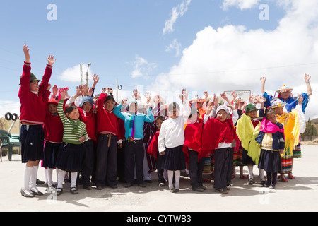 A group of children, likely students, are standing together outdoors on a paved surface under a bright blue sky with scattered clouds. They are dressed in a mix of school uniforms—red sweaters, navy Stock Photo