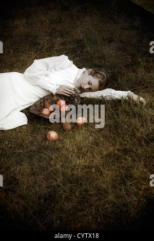 a woman in a white edwardian dress is lying in the grass, in front of her a basket with fruits Stock Photo