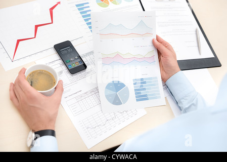 Businessman at the workplace drink coffee and looking some papers with charts and numbers. Stock Photo