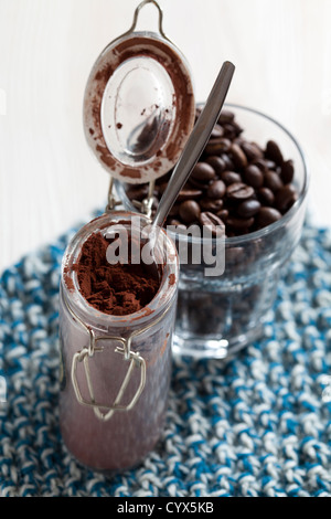 Close up of a glass jar of cocoa powder with open lid and spoon with coffee beans in background Stock Photo