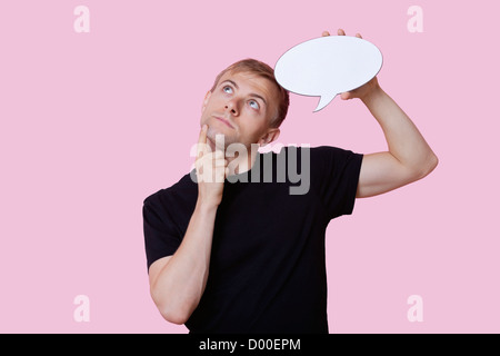 Thoughtful young man holding speech bubble while looking up over pink background Stock Photo