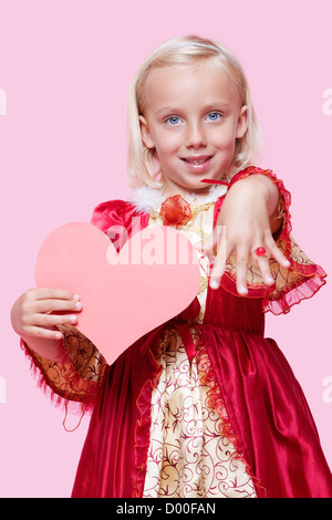 Portrait of a happy young girl dressed in princess costume holding paper heart as she displays ring over pink background