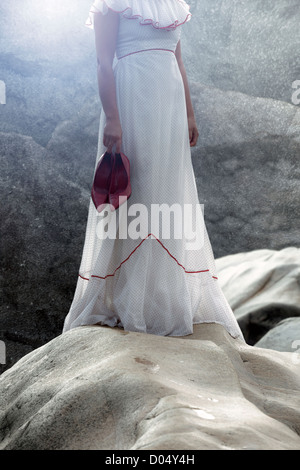 a woman in a white dress with polka dots is standing on rocks, holding her shoes in her hands Stock Photo