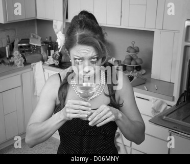 Weeping Caucasian woman drinks a martini in her messy kitchen Stock Photo