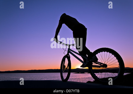 A trials bike rider balances on a wall at sunset with a seascape and low hills in the background - No Sales on 17�Թ����� or other Stock Photo