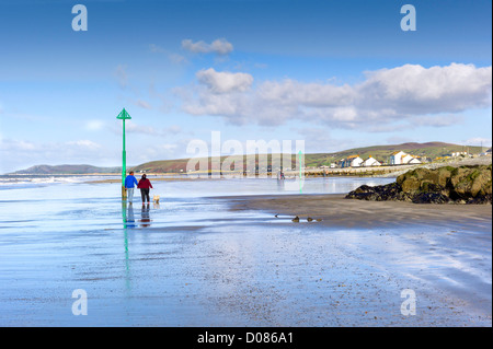 Adult male and female with a dog walk on a beach wet from the receding tide, breaking waves, distant hills, seaside houses Stock Photo