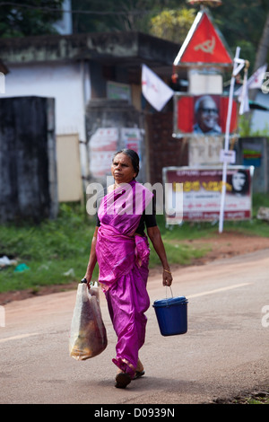 INDIAN WOMAN RETURNING FROM MARKET PROPAGANDA POSTER FROM KERALA COMMUNIST PARTY ON STREET IN NEDUNGOLAM KERALA SOUTHERN INDIA Stock Photo