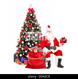 Santa Claus with a gift sitting next to a bag full of presents, christmas tree in the background Stock Photo