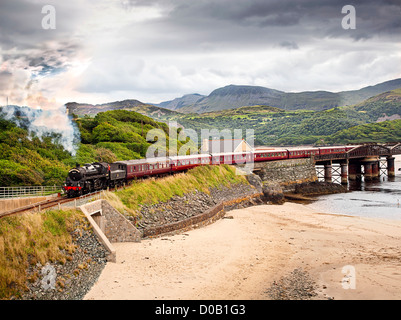 Steam train the Cambrian crosses Barmouth rail bridge over the Mawddach estuary with mountains in the background, near beach. Stock Photo