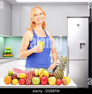 Woman holding a glass of orange juice in front of pile of fruit with kitchen in the background Stock Photo