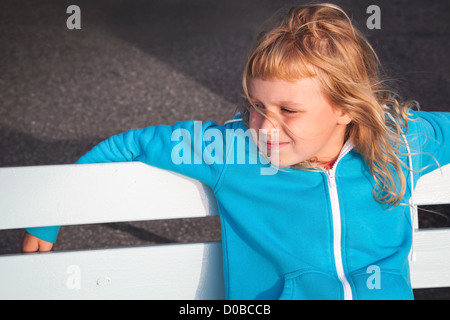 Little blond girl in casual sport outwear relaxes with smile on white park bench Stock Photo