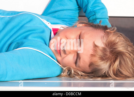 Little blond girl in casual sport outwear lies and relaxes with smile on white park bench Stock Photo