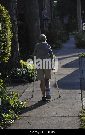 A woman with an injured right foot walks cautiously on two crutches, outdoors, on a pavement, Ontario, Canada.  Vertical. Copy space. Rehab. Stock Photo