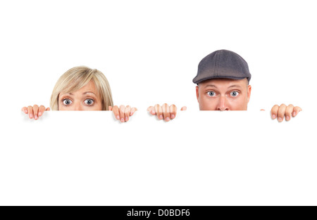 Young couple, man and woman humorous faces with big eyes, looking out from behind a blank white sign with funny expression. Stock Photo