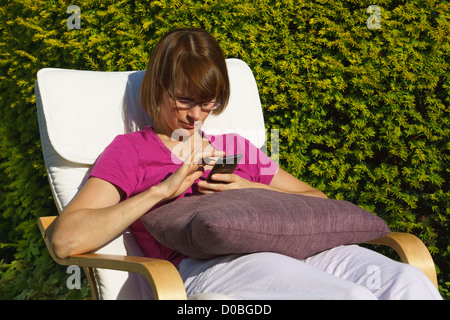 A young woman checking her mobile phone while sitting in a sunny garden. Stock Photo