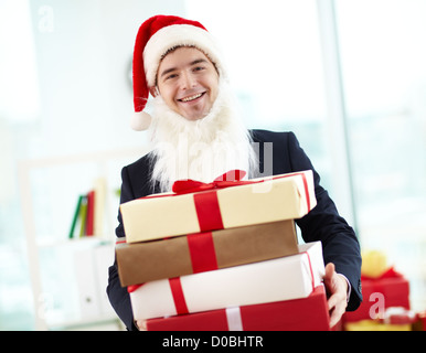 Image of happy businessman in Santa cap and beard holding stak of gifts and looking at camera in office Stock Photo
