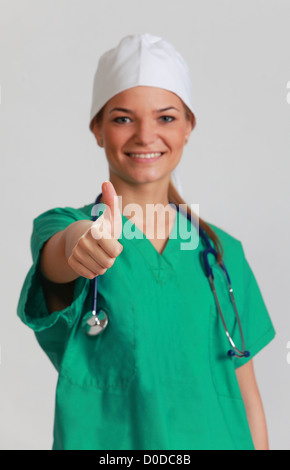Portrait of a smiling young woman doctor with her right thumb up, against a gray background. Selective focus on the thumb. Stock Photo