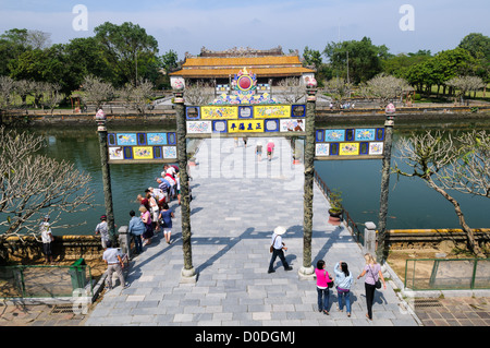 Imperial City Main Entrance Bridge Ceramic Mosaic Gate Hue Vietnam | HUE, Vietnam — Visitors cross a bridge through a ceramic mosaic gate inside the Imperial City, moving toward Thai Hoa Palace in the background. The central path was historically reserved exclusively for the Nguyen emperors; court officials approached by flanking routes. The palace's yellow glazed roof tiles — a color strictly restricted to the sovereign — marked the political heart of the dynasty from its founding in 1802 until Bao Dai's abdication in 1945. Stock Photo