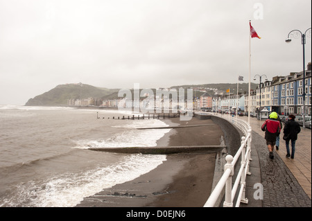 ABERYSTWYTH, Wales — A handful of pedestrians brave blustery and rainy conditions as they walk along the waterfront promenade in Aberystwyth, a coastal town on the western shore of Wales. The stormy weather over Cardigan Bay creates a dramatic scene along this popular seaside destination, showcasing the raw beauty of the Welsh coast. Stock Photo