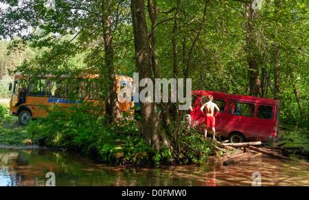 Yellow school bus tows red minibus from broken wooden bridge. Stock Photo