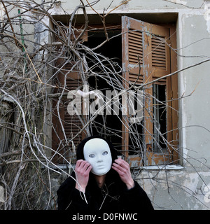 Grotesque white mask figure and haunted house window with tangled overgrown plants and creepy dummy doll hand. Stock Photo