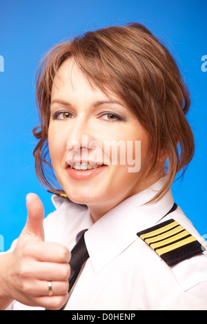 Beautiful airline pilot wearing uniform with epauletes showing thumb up gesture of approval, standing on blue background. Stock Photo