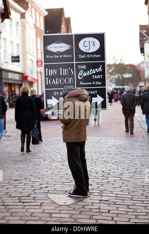 A man stood in the high street holding a placard with directions for local shops. Stock Photo