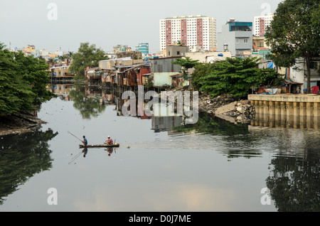 HO CHI MINH CITY, Vietnam - Two men in a small wooden canoe use a net to fish in the still waters of a tributary of the Saigon River in Ho Chi Minh City, Hanoi, with makeshift houses on the waterfront and tall highrises in the background. Stock Photo