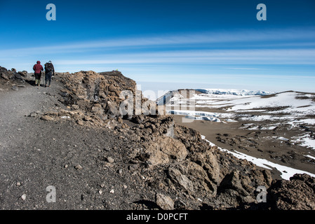 Mount Kilimanjaro Hikers Approaching Uhuru Peak Tanzania | MOUNT KILIMANJARO, Tanzania — Two hikers pause on a rocky ridge above Kilimanjaro's equatorial glaciers, with the signboard at Uhuru Peak, the mountain's 19,341-foot (5,895 m) summit, visible on the skyline beyond. The exposed ice fields at right sit on Kibo, the dormant cone forming the highest of the mountain's three volcanic centers, and are projected to vanish within decades. Stock Photo