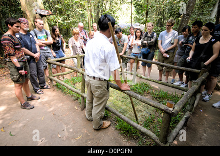 HO CHI MINH CITY, Vietnam — A tour guide demonstrates a preserved punji trap at the Cu Chi Tunnels historical site, showing visitors one of many defensive measures used during the Vietnam War. The camouflaged trap features a concealed pit with metal spikes, a common defensive technique employed throughout the conflict. The demonstration is part of the site's educational exhibits showcasing Vietnamese military tactics. Stock Photo