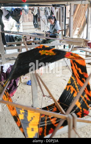 A woman using her loom on the dirt floor under her house in northeastern Laos. At right, in the foreground, is some of the brightly dyed silk thread she's using, and at bottom right you can see part of a bicycle wheel that has been repurposed for part of the spindle. Stock Photo