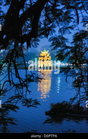 Turtle Tower (also known as Tortoise Tower on a small island in Hoan Kiem Lake in the historical center of Hanoi, Vietnam. The lights of the tower are on and reflected on the water, with the image framed by the branches of a tree on the shore. Stock Photo