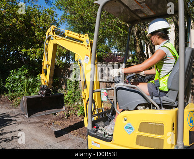 A young woman construction worker, working using a digger excavator machine wearing protective white hard hat and high vis vest. Stock Photo