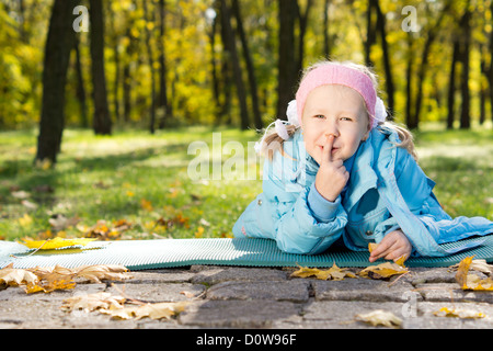 Smiling little girl lying on a mat in the park making shushing gesture raising her finger to her lips as a sign of silence or se Stock Photo