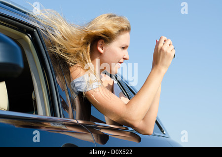 Young woman with her blonde hair blowing in the breeze leaning out of a car taking photographs Stock Photo