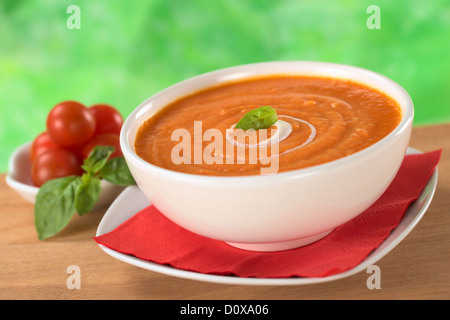 Cream of tomato with a small spiral of cream on top and a basil leaf with tomatoes and basil leaves in the back Stock Photo