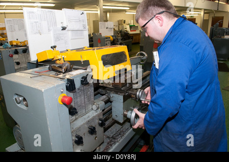 Young white man in blue overalls on a factory floor on an engineering apprenticeship wearing protective safety glasses operating a machine Stock Photo