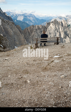 Lone man sits on bench seat at edge of Austrian mountain peak, looking out over the Alps with layers of rugged rocky mountains. Stock Photo