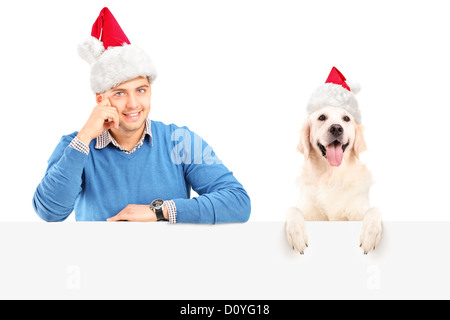 Smiling guy and dog wearing santa claus hats and posing behind a blank panel Stock Photo