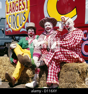 1970s THREE LAUGHING CLOWNS SITTING ON HAY BALES BY CIRCUS WAGON LOOKING AT CAMERA Stock Photo