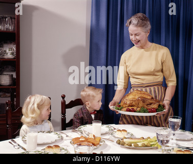 1960s GRANDMOTHER PRESENTING ROASTED TURKEY TO CHILDREN AT DINING ROOM TABLE Stock Photo