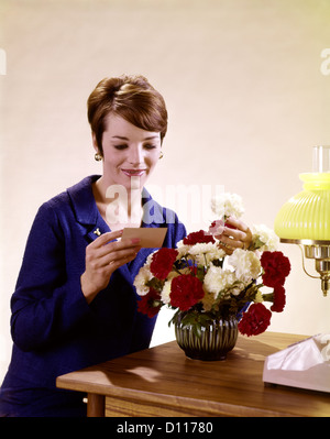 1960s WOMAN READING GIFT CARD WITH A VASE RED WHITE CARNATIONS Stock Photo