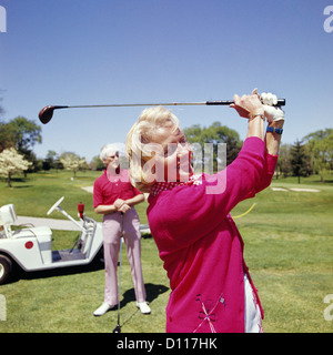 1960s MATURE WOMAN GOLFING AS MAN STANDS BY GOLF CART Stock Photo