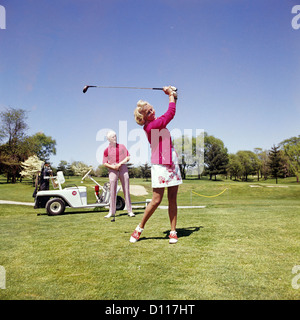1960s 1970s MATURE WOMAN SWINGING GOLF CLUB AS MAN STANDS BY GOLF CART Stock Photo