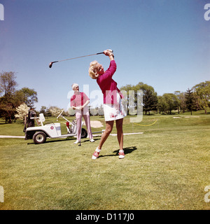 1970s MATURE WOMAN SWINGING GOLF CLUB AS MAN STANDS BY GOLF CART Stock Photo