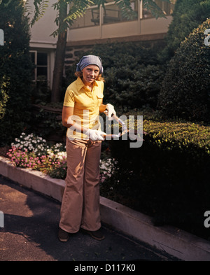 1970s WOMAN USING GARDEN CLIPPERS TRIMMING BOXWOOD HEDGE WEARING BELLBOTTOMS Stock Photo